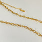 A gold chain necklace with elongated oval links and a small gold tag, displayed on a textured off-white background.