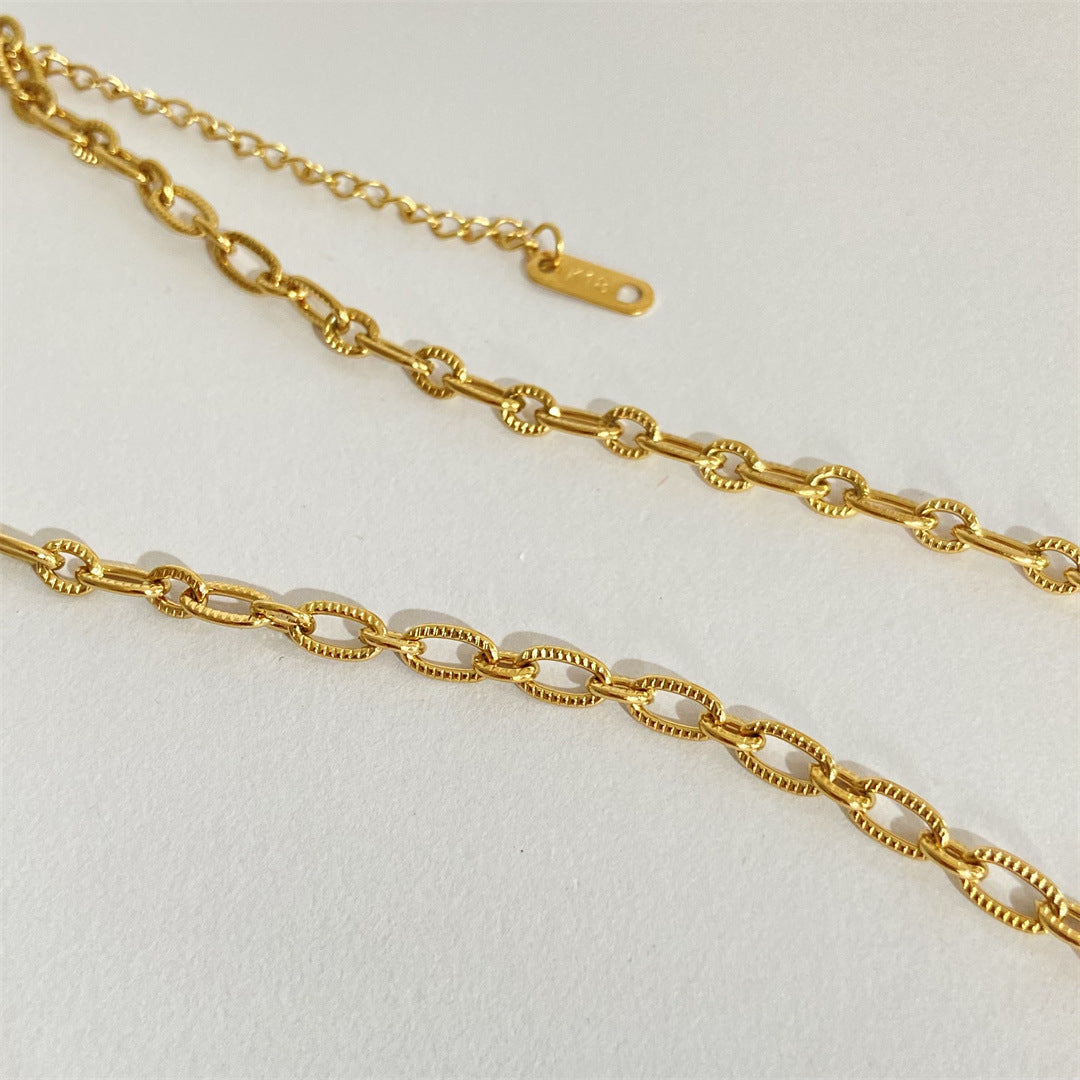 A gold chain necklace with elongated oval links and a small gold tag, displayed on a textured off-white background.