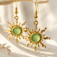 A pair of gold-toned dangle earrings with sunburst-shaped metal arms and oval light green cabochon stones, hanging from a curved cream branch against a bright white background with soft shadows.