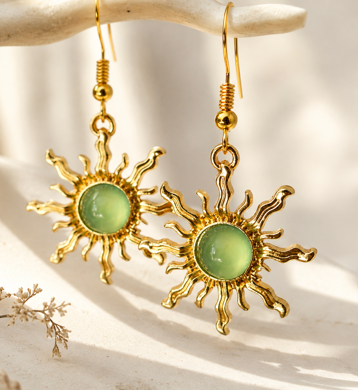 A pair of gold-toned dangle earrings with sunburst-shaped metal arms and oval light green cabochon stones, hanging from a curved cream branch against a bright white background with soft shadows.