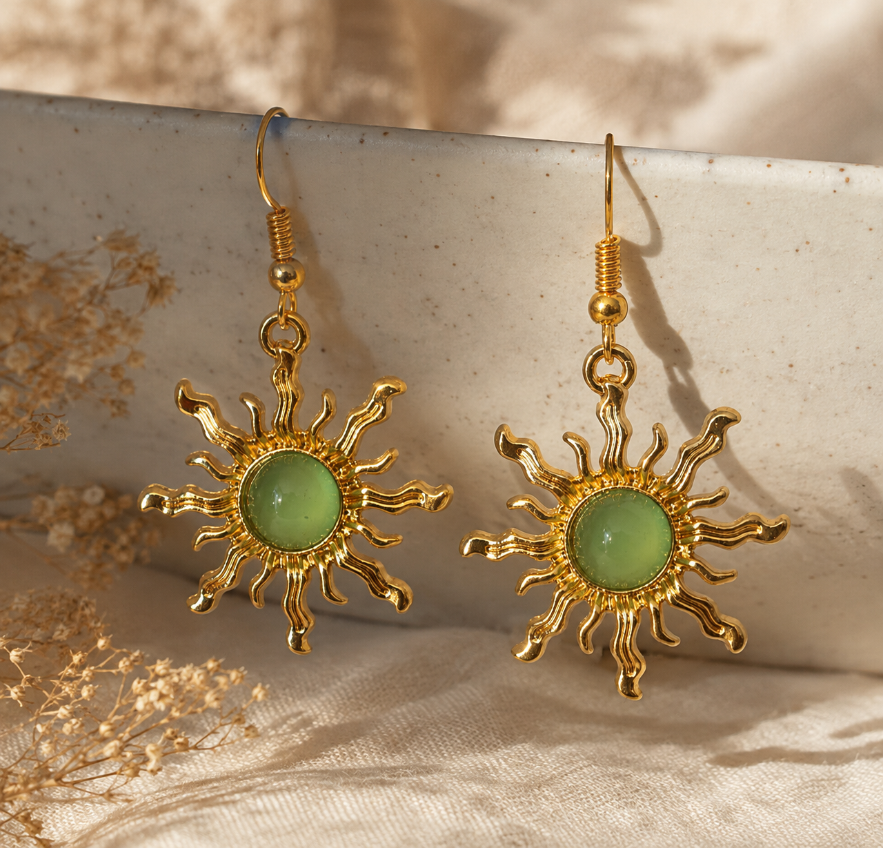 A pair of gold sunburst drop earrings with oval light green cabochon stones, displayed on a textured ceramic dish with dried floral elements.