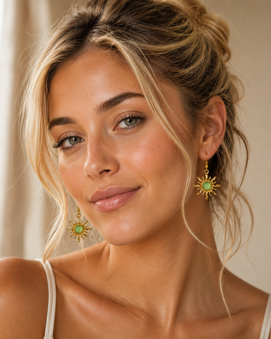 A close-up portrait of a woman wearing gold sunburst dangle earrings with green oval centers, set against a soft beige background.