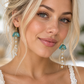 A close-up of a blonde woman wearing dangling earrings with turquoise blue bell-shaped tops and clear bead drops, paired with a white lace top.