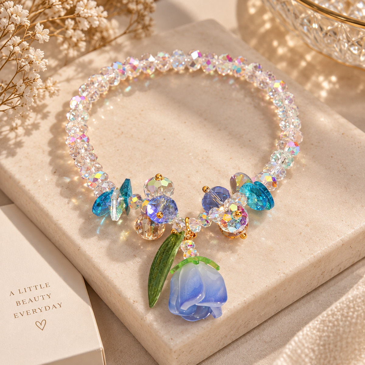 A beaded crystal necklace with blue and iridescent faceted beads, gold-tone accents, and a dangling blue rose charm with green leaves, displayed on a cream stone slab with dried flowers and a small card.