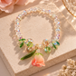A beaded bracelet with iridescent clear beads, green crystal leaves, and a dangling peach rose charm, displayed on a beige stone slab with warm sunlight and soft floral props.