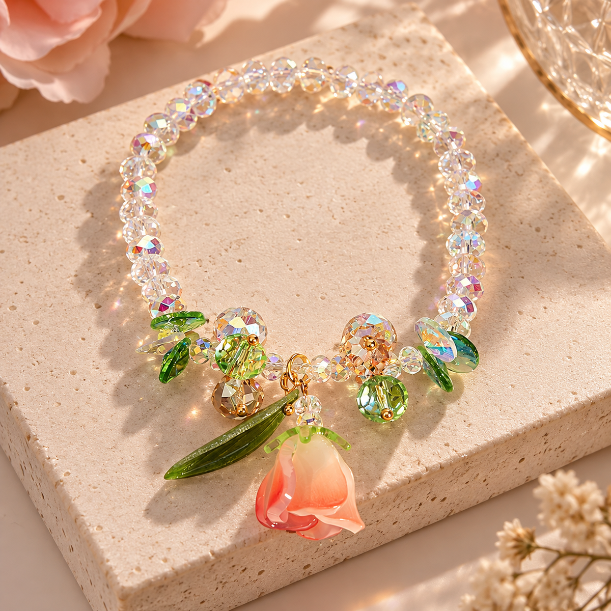 A beaded bracelet with iridescent clear beads, green crystal leaves, and a dangling peach rose charm, displayed on a beige stone slab with warm sunlight and soft floral props.