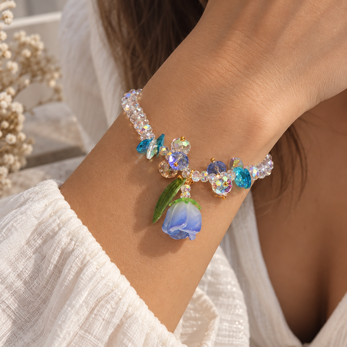 A close-up of a person's wrist wearing a beaded charm bracelet. The bracelet features iridescent clear rhinestones, blue faceted beads, a small gold-toned heart, and a dangling blue rose charm with green leaves.