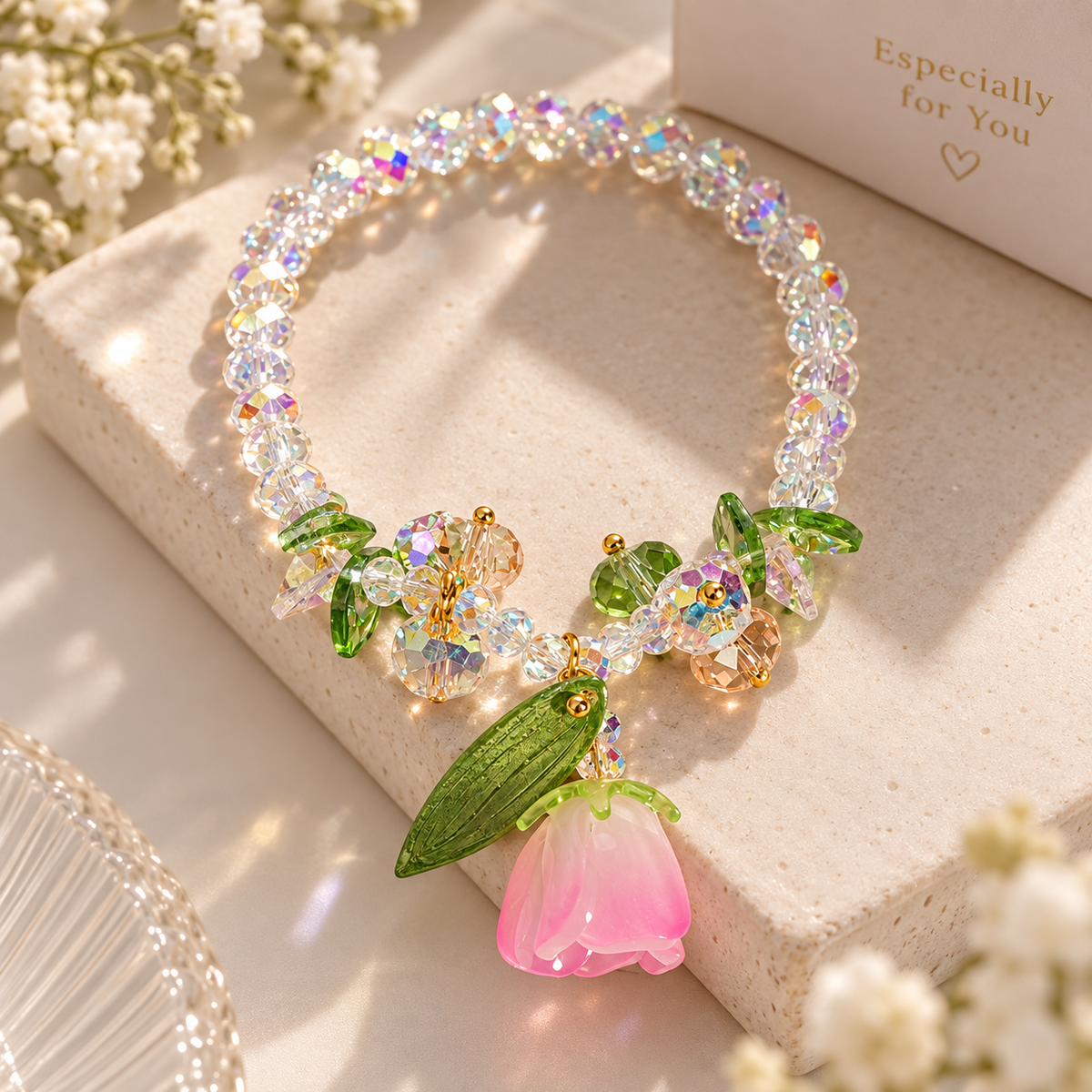A floral beaded necklace with a pink bell-shaped flower charm, displayed on a textured beige stone block with decorative white flowers and a card in the background.
