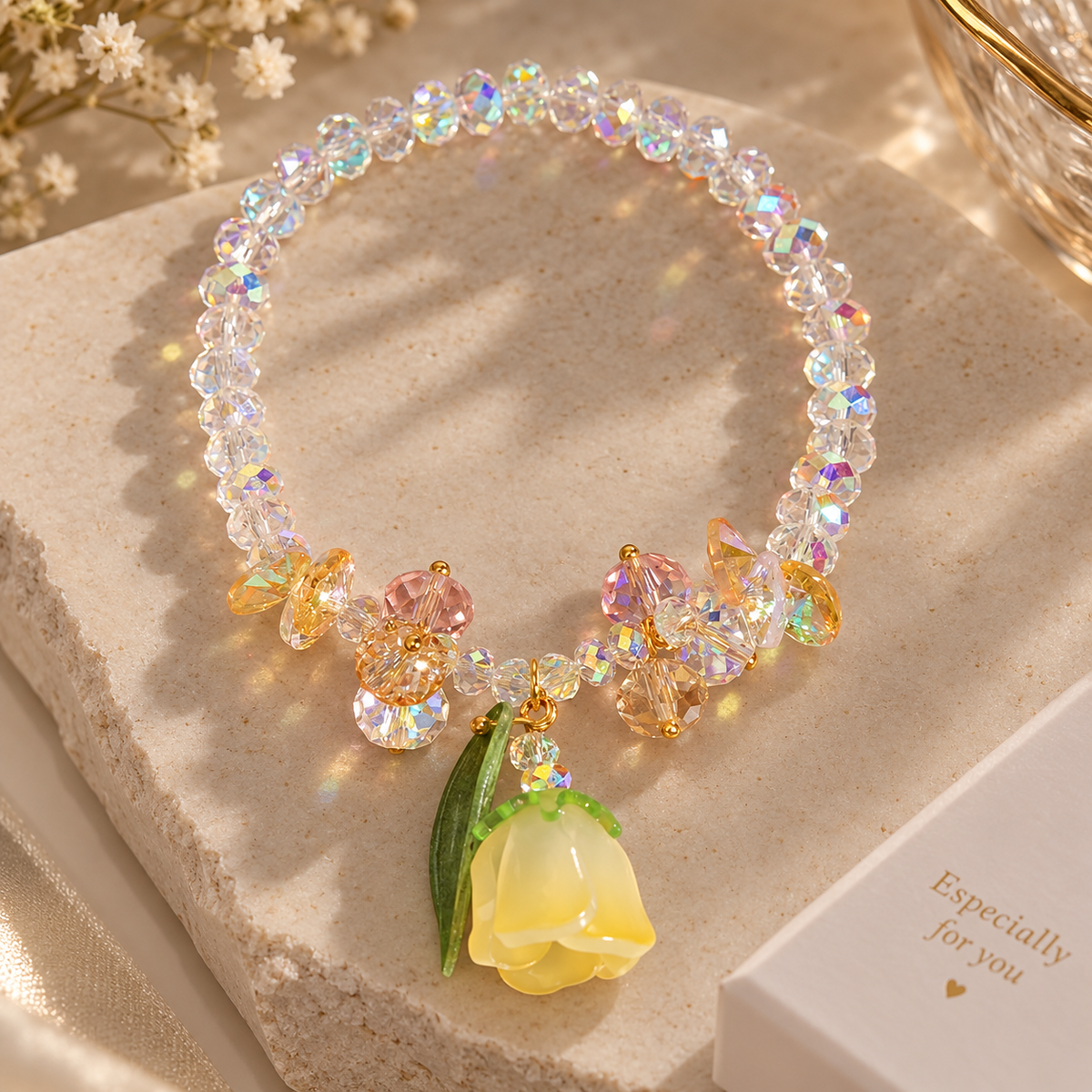 A beaded bracelet with iridescent clear beads, gold-tone accents, and a yellow flower charm with green leaves, displayed on a textured beige stone block.