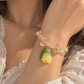 A close-up of a person's wrist wearing a delicate bracelet with iridescent crystal-like beads, a yellow flower charm, and green leaves, styled with a white lace top.