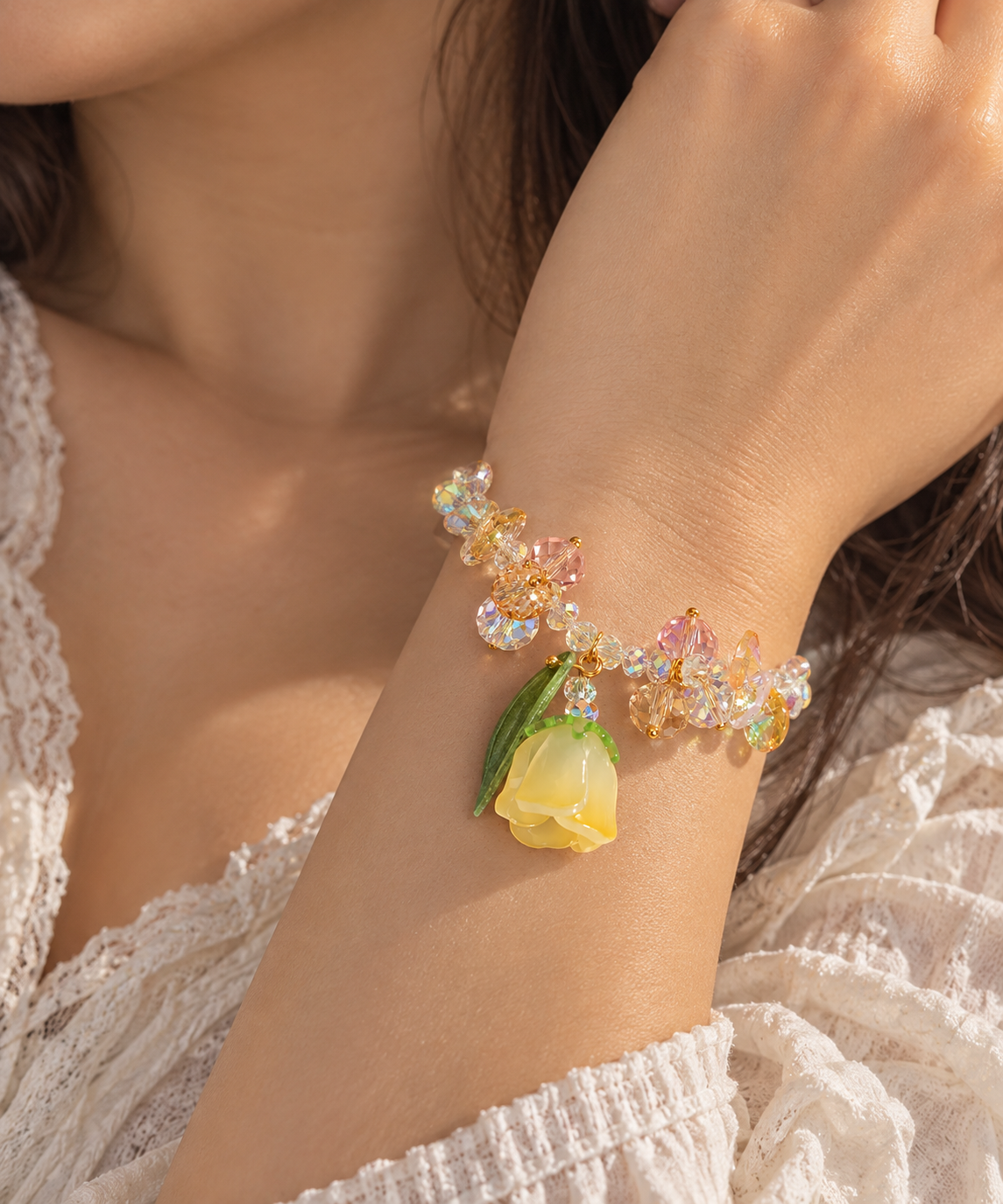 A close-up of a person's wrist wearing a delicate bracelet with iridescent crystal-like beads, a yellow flower charm, and green leaves, styled with a white lace top.