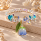 A beaded crystal bracelet with iridescent and blue faceted beads, gold-tone connectors, and a dangling flower charm in blue and green, displayed on a beige stone surface with soft decorative flowers in the background.