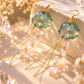 A pair of gold-tone dangle earrings with round pale green beads and clear crystal drops, displayed on a warm beige surface with soft floral props and sparkling light effects.