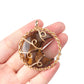 Hand holding a gold-framed pendant with a brown stone on a white background