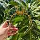 A gold metal headband decorated with green stone beads and a gold crescent moon centerpiece, held by a hand against a background of large green monstera leaves.