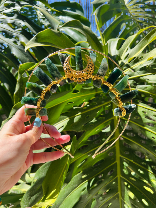 A gold metal headband decorated with green stone beads and a gold crescent moon centerpiece, held by a hand against a background of large green monstera leaves.