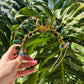 A gold crescent moon and star filigree crown decorated with green stone beads, held by a hand against a background of large green monstera leaves.