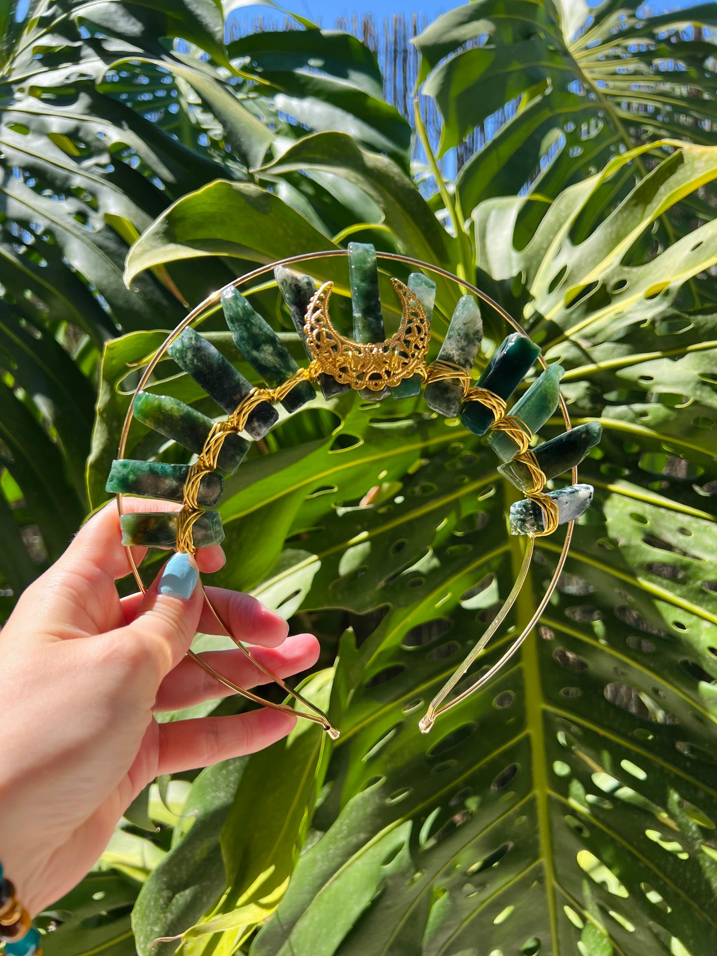 A gold crescent moon and star filigree crown decorated with green stone beads, held by a hand against a background of large green monstera leaves.