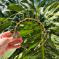 A gold wire headband decorated with green stone beads, held by a hand against a background of large green monstera leaves.