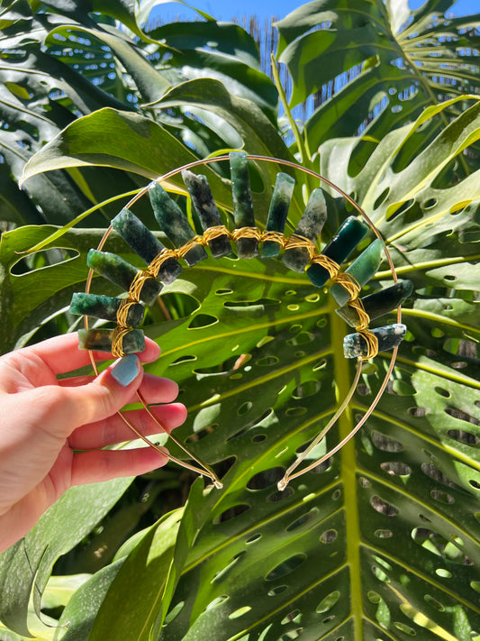 A gold wire headband decorated with green stone beads, held by a hand against a background of large green monstera leaves.