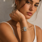 A close-up lifestyle shot of a woman wearing a gold-toned three-band cuff bracelet with a central light blue rectangular stone cluster, set against a warm, sunlit neutral background.