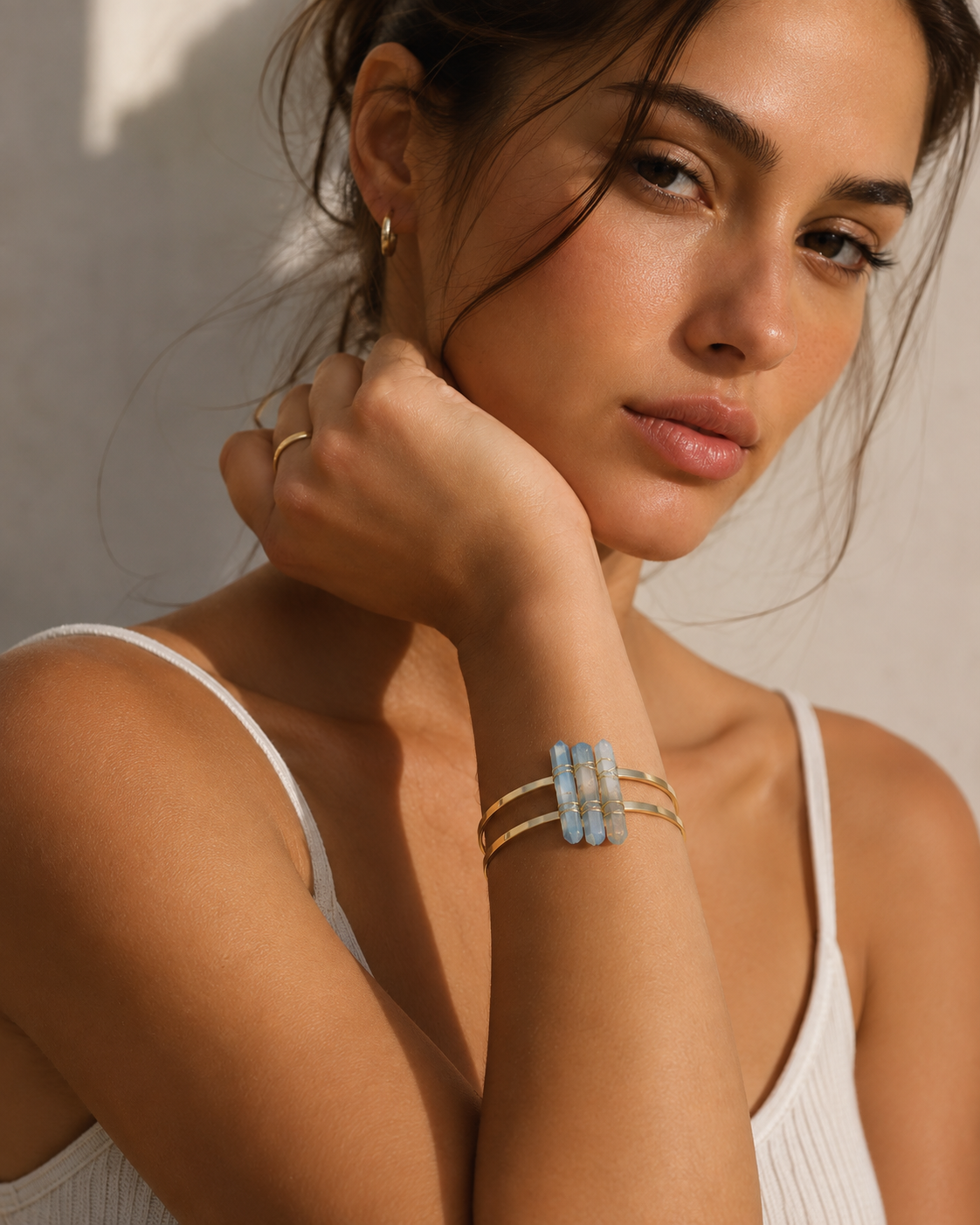 A close-up lifestyle shot of a woman wearing a gold-toned three-band cuff bracelet with a central light blue rectangular stone cluster, set against a warm, sunlit neutral background.