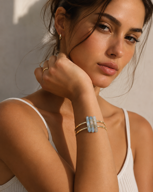 A close-up lifestyle shot of a woman wearing a gold-toned three-band cuff bracelet with a central light blue rectangular stone cluster, set against a warm, sunlit neutral background.