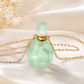 A light green faceted gemstone perfume bottle with a gold chain and gold cap, displayed on draped white satin fabric with dried flowers in the background.