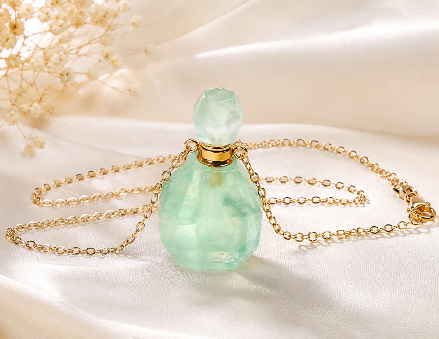 A light green faceted gemstone perfume bottle with a gold chain and gold cap, displayed on draped white satin fabric with dried flowers in the background.