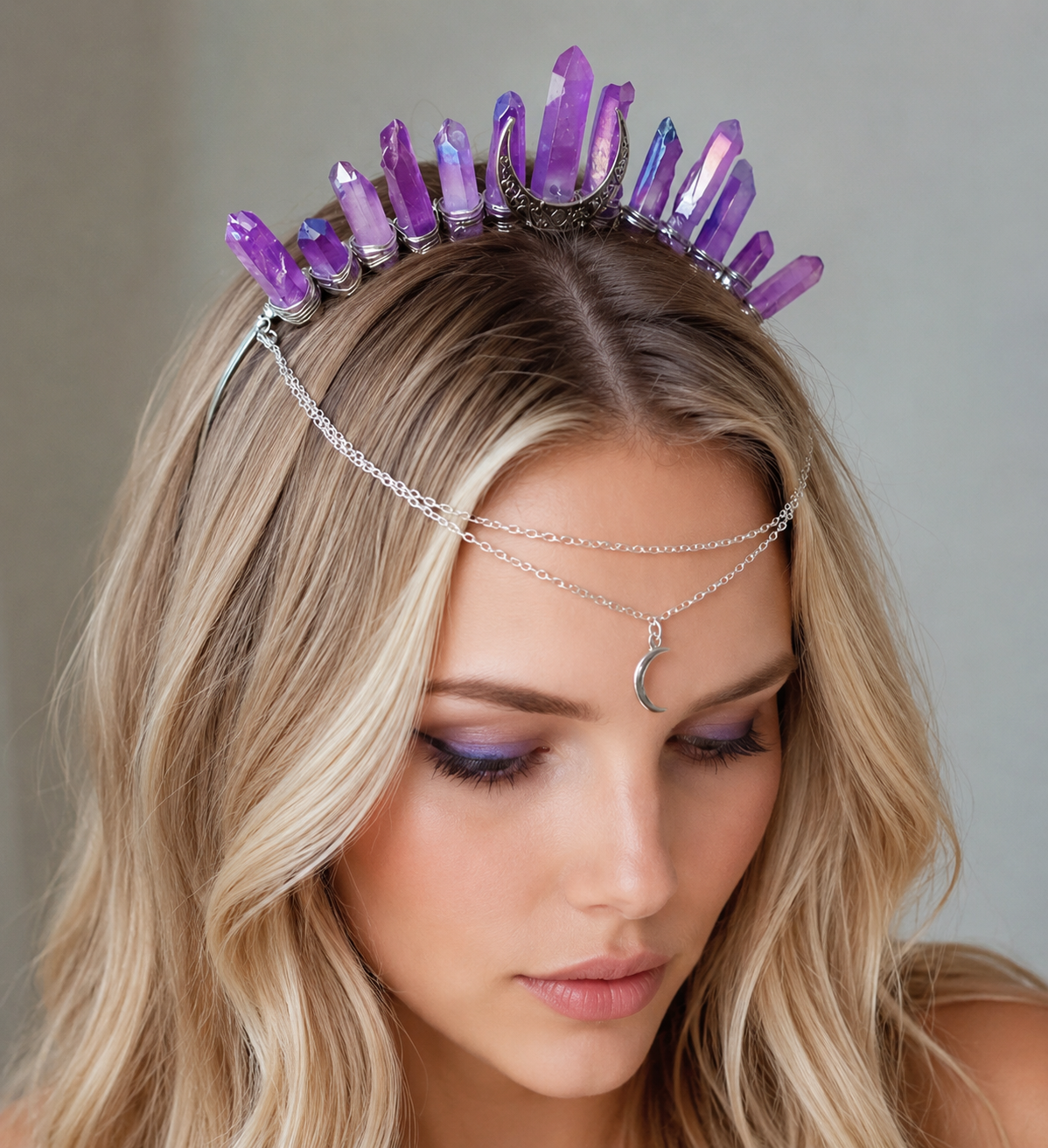 A woman with blonde hair wearing a purple crystal tiara and a silver chain headpiece with a crescent moon pendant, looking down against a neutral gray background.