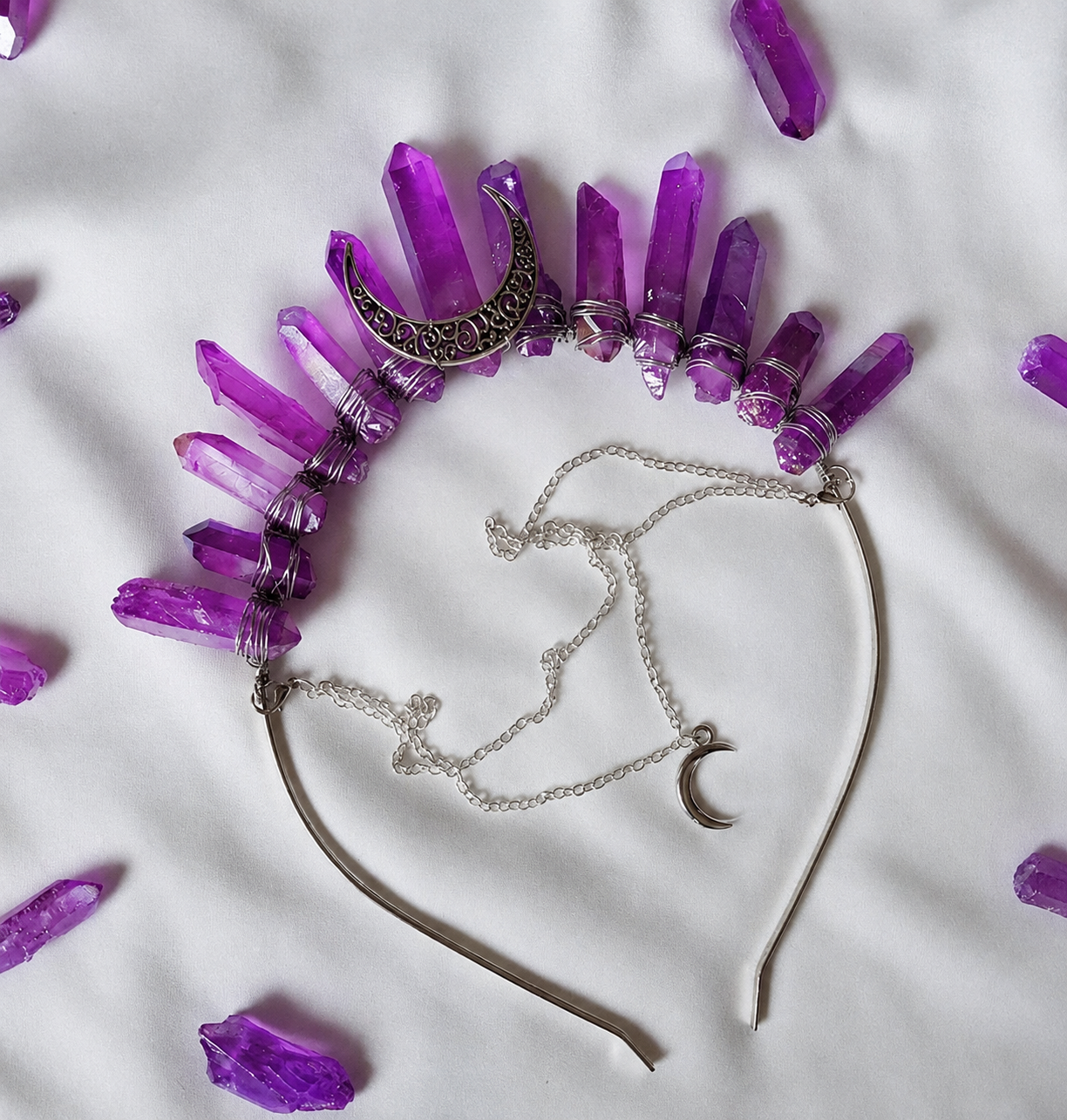 A headband necklace featuring multiple purple amethyst crystal points and a silver crescent moon charm, laid flat on a white fabric background with scattered loose crystals.