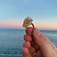 A handmade gold wire ring with a raw, organic-shaped stone held up by a hand against a blurred beach sunset background.