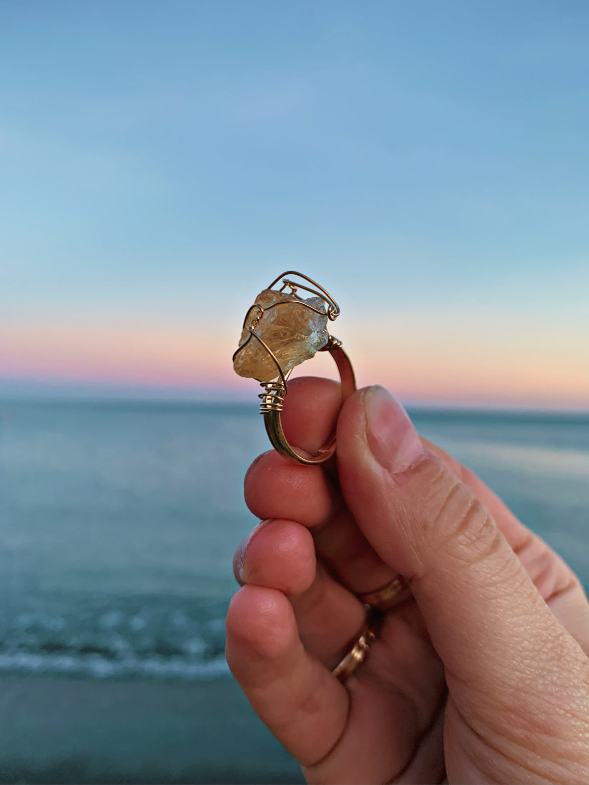 A handmade gold wire ring with a raw, organic-shaped stone held up by a hand against a blurred beach sunset background.
