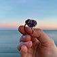 A hand holding a gold wire-wrapped ring with a deep purple, textured stone, set against a blurred ocean and sunset background.