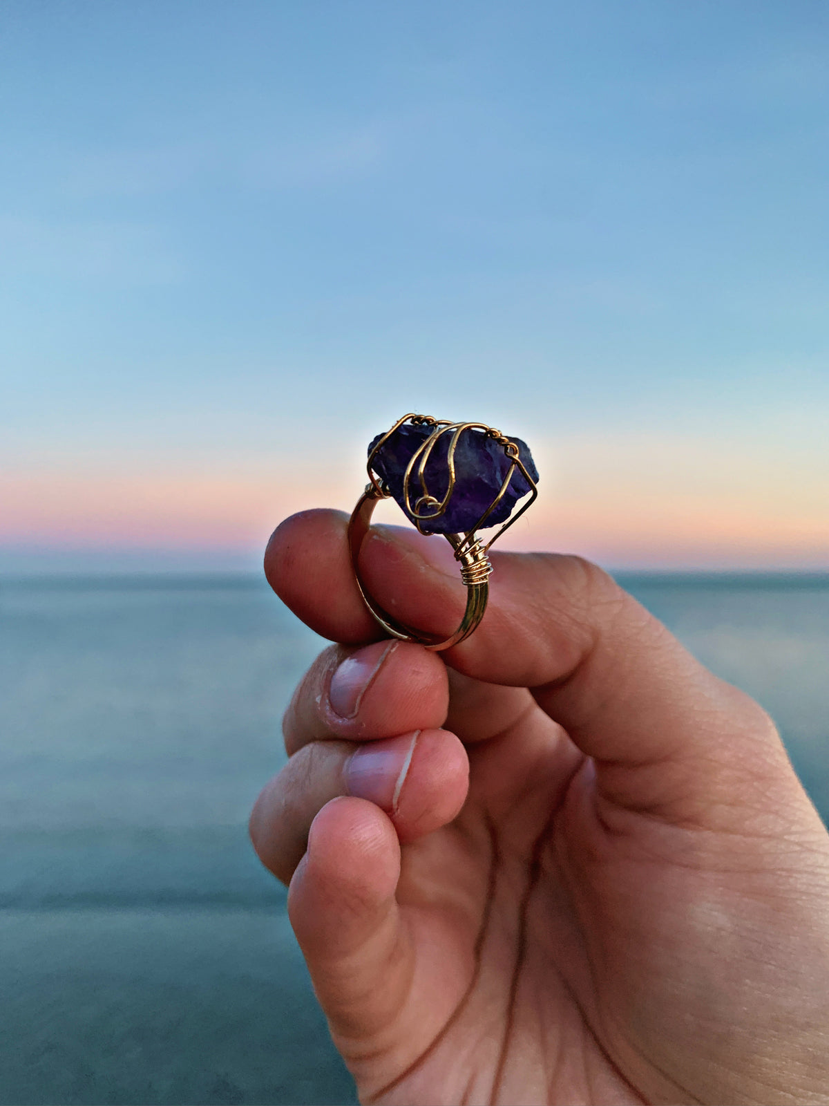 A hand holding a gold wire-wrapped ring with a deep purple, textured stone, set against a blurred ocean and sunset background.
