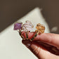 A close-up of a hand holding three raw gemstone wire-wrapped rings. The rings feature purple amethyst, pale pink quartz, and golden citrine stones in gold-toned wire settings, photographed against a warm, softly lit background.