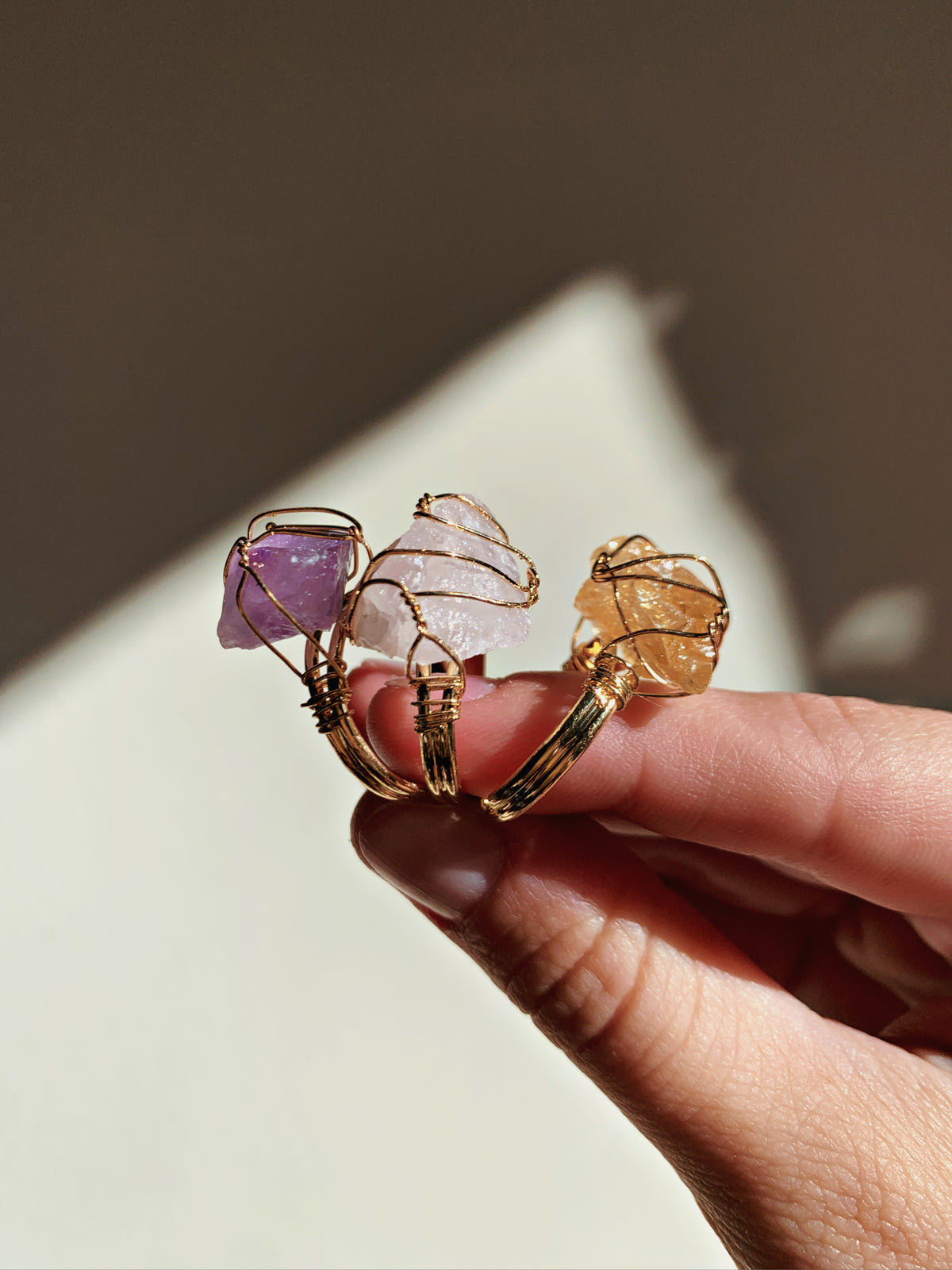 A close-up of a hand holding three raw gemstone wire-wrapped rings. The rings feature purple amethyst, pale pink quartz, and golden citrine stones in gold-toned wire settings, photographed against a warm, softly lit background.