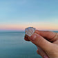 A hand holding a small sea glass ring with a milky white stone and gold wire details, set against a blurred ocean and sunset background.