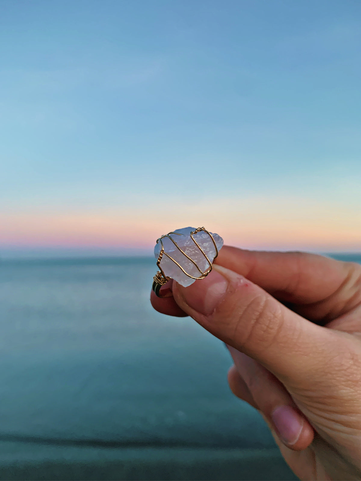 A hand holding a small sea glass ring with a milky white stone and gold wire details, set against a blurred ocean and sunset background.