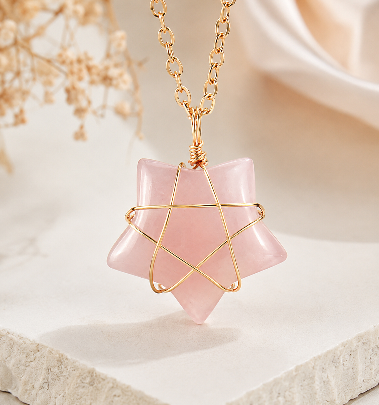 A pink rose quartz star pendant necklace with a gold wire star design wrapped around the center, hanging from a gold link chain on a soft, light beige background.