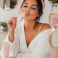 Woman in a white robe with a pink headband, surrounded by skincare products in a cozy room.