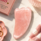Pink gua sha tool on a marble surface with 'Self Love Club' book and jewelry.