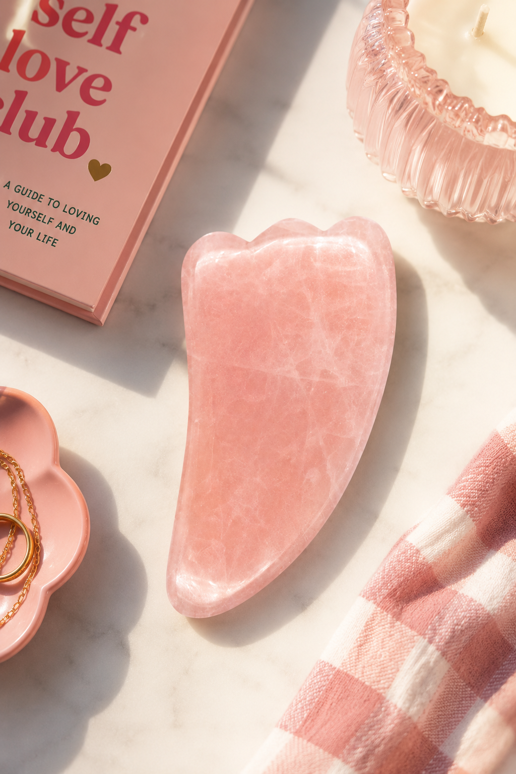 Pink gua sha tool on a marble surface with 'Self Love Club' book and jewelry.