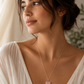 A woman wearing a delicate gold chain necklace with a pink stone pendant, styled with a white draped top against a soft, warm neutral background.
