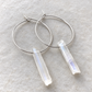 Silver hoop earrings with rectangular milky white crystal pendants, displayed on a textured light gray stone surface.