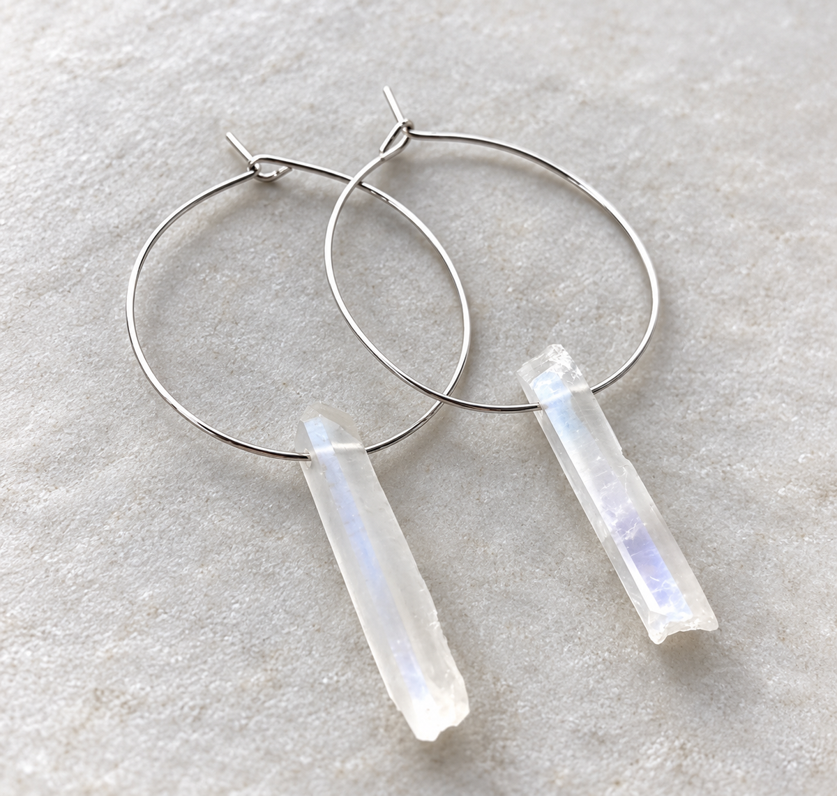 Silver hoop earrings with rectangular milky white crystal pendants, displayed on a textured light gray stone surface.