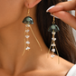 A close-up lifestyle shot of a person wearing and holding a pair of gold dangle earrings with dark green jellyfish-shaped tops and clear faceted bead tails.