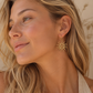 A close-up of a woman with blonde hair wearing a gold sunburst dangle earring, set against a warm beige background.