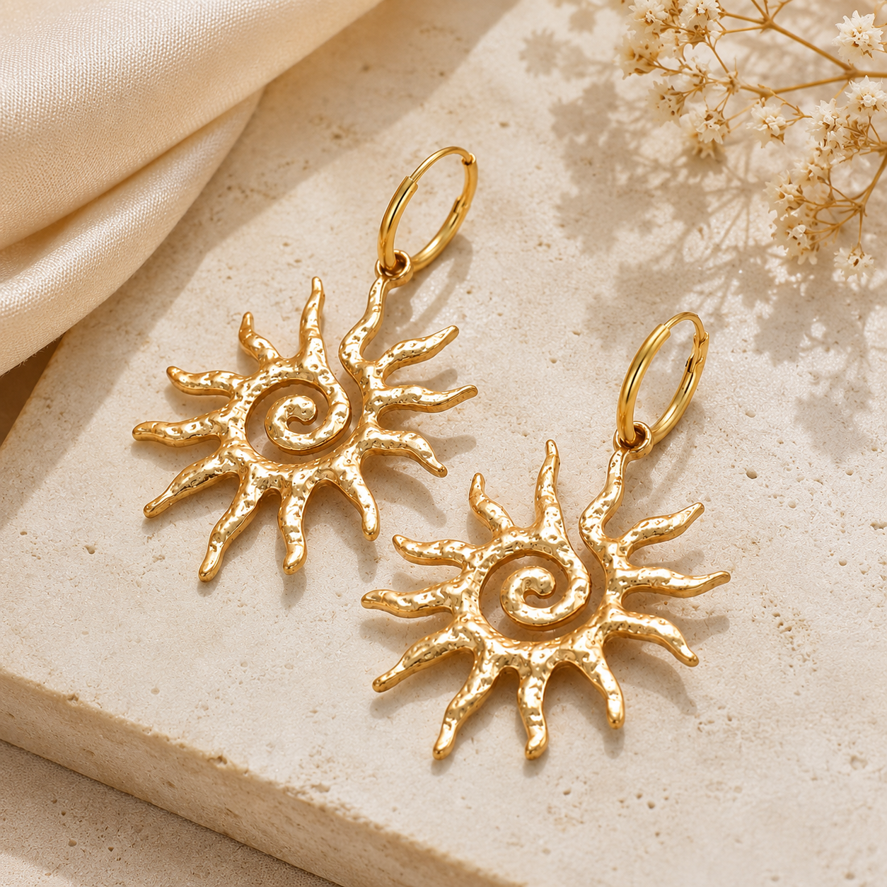 A pair of gold-toned sunburst dangle earrings with textured spiral centers, displayed on a light beige stone surface with cream fabric and dried flowers.