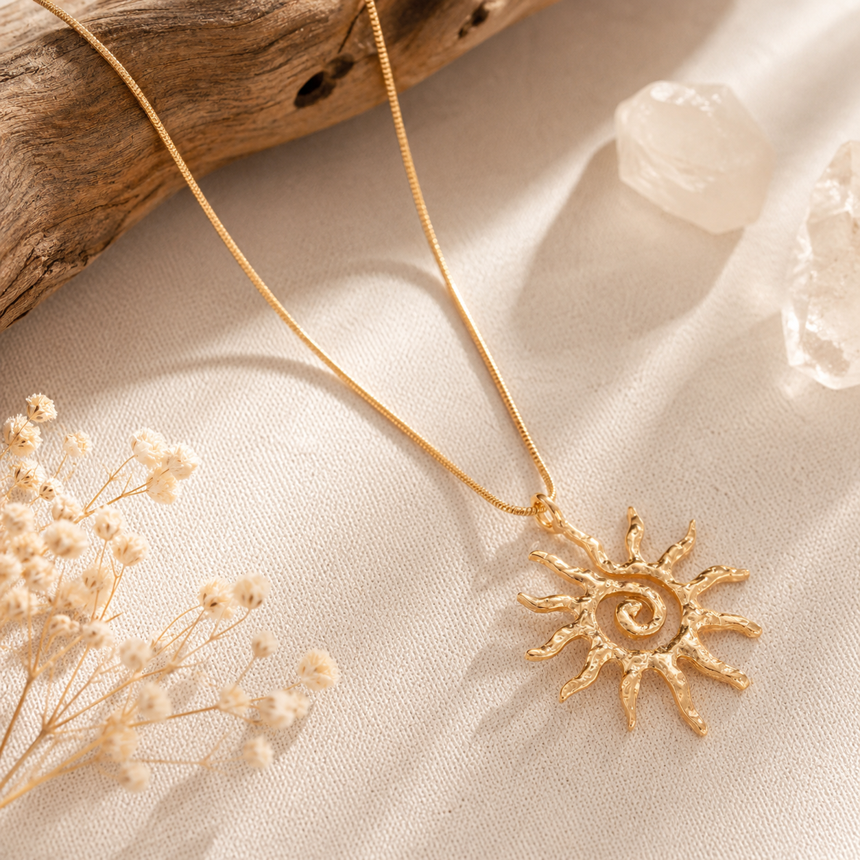 A gold-toned sun pendant necklace with a textured spiral center, resting on a beige fabric surface with dried flowers and a piece of driftwood.
